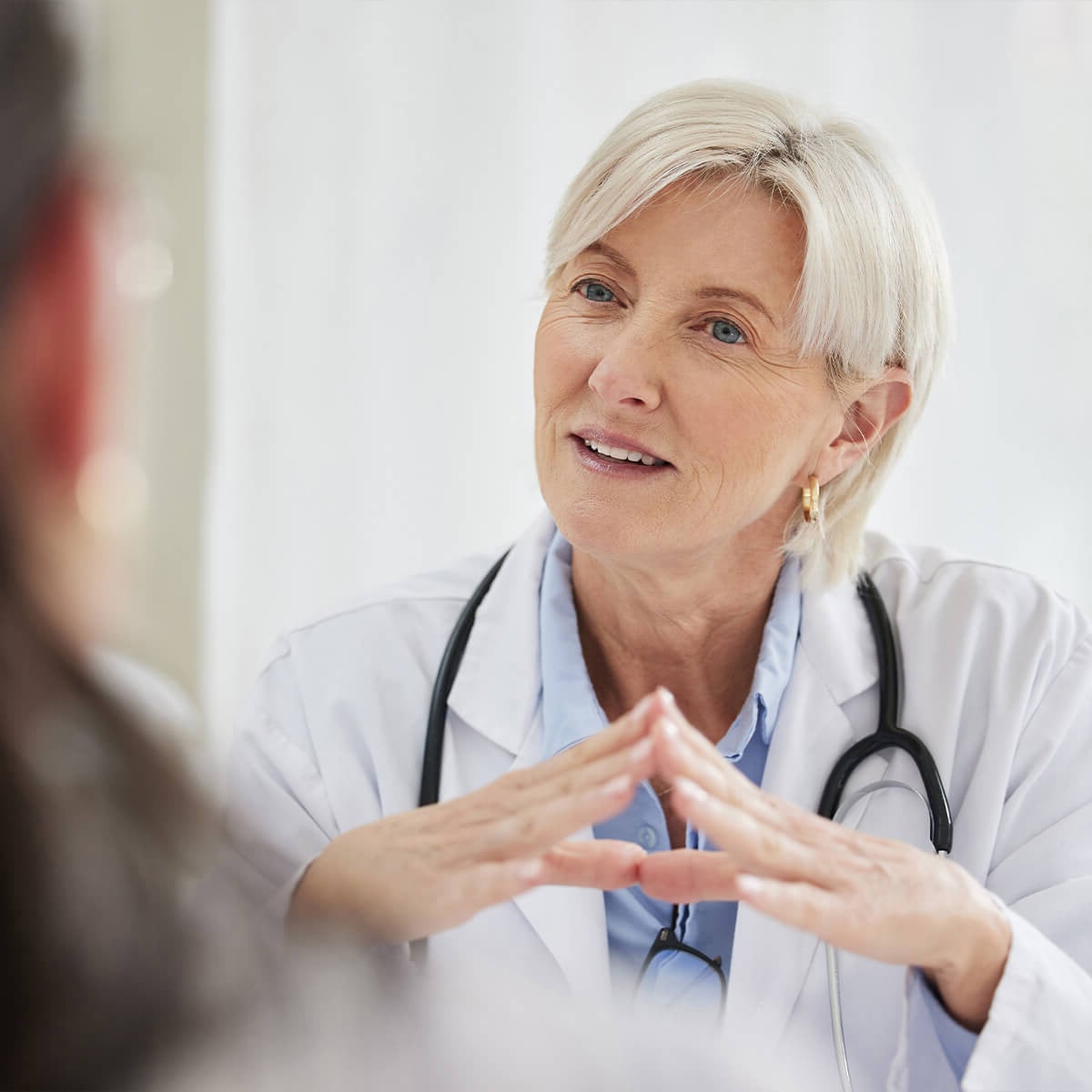 Eine Ärztin, die mit gefalteten Händen dasitzt, einen weißen Kittel und ein Stethoskop trägt und sich in einer medizinischen Umgebung mit einem Patienten unterhält.